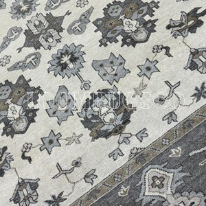 Puede incluir: Primer plano de una alfombra beige y gris estampada. La alfombra presenta un diseño floral y geométrico repetido en tonos grises, marrones y beige. El borde es de un gris más oscuro con patrones similares. La alfombra tiene una estética vintage o antigua.