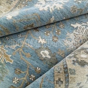 May include: Close-up of a rolled-up area rug with a distressed, vintage-inspired design. The rug features a light blue background with floral and geometric patterns in shades of beige, brown, and white. The text "LOOMRUGSCO" is visible.