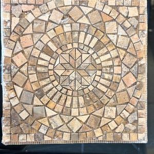May include: Square mosaic tile with a circular pattern in shades of brown, beige, and tan. The design features a central star shape surrounded by concentric rings of irregularly shaped tiles. The tile is made of natural stone.