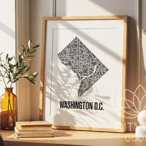 May include: A framed print of Washington D.C. features a black and white map of the city, composed of neighborhood names. The print is in a light wood frame and is displayed on a wooden surface. The words "WASHINGTON D.C." are printed below the map.
