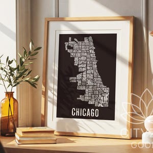 May include: A framed art print featuring a black silhouette of Chicago filled with white text of city neighborhoods. The word "CHICAGO" is at the bottom. The print is in a light wood frame, displayed on a wooden surface with a vase and books.