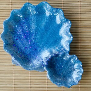 May include: A blue resin dish with a wavy, scalloped edge. The dish has a smaller, attached compartment. Glitter is embedded in the blue resin, creating a sparkling effect. The dish is on a light brown bamboo mat.