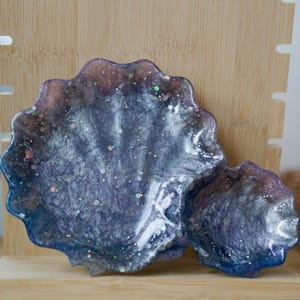 May include: A decorative resin tray with a unique, organic shape. The tray features a swirling mix of dark blue, purple, and silver colors, with embedded glitter for added sparkle. The tray is approximately 6 inches wide and 4 inches long.