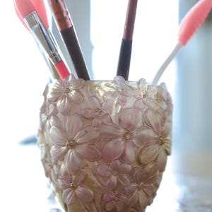 May include: A decorative makeup brush holder with a floral design. The holder is filled with various makeup brushes and applicators. The holder is a light pink color with gold accents.