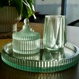 Könnte beinhalten: Ein dekoratives Set aus Glas-Wohnaccessoires. Das Set umfasst ein rundes Tablett, ein Glas mit Deckel und ein facettiertes Glas. Alle Artikel sind hellgrün und stehen auf einer Holzoberfläche.