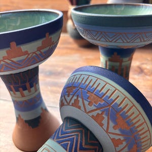 May include: Three ceramic hookah bowls featuring a matte finish with geometric patterns in shades of blue, green, and terracotta. Each bowl has a wide rim, a tapered neck, and a flared base, decorated with intricate zig-zag and block designs.