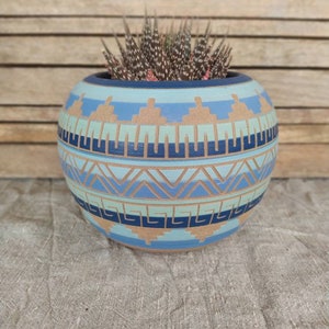 Può includere: Un vaso rotondo in ceramica dipinto a mano con un design geometrico nei toni del blu, verde acqua e beige. Il vaso è riempito con una piccola pianta succulenta. Il design presenta motivi ripetuti di triangoli e forme rettangolari. Il vaso misura circa 15 cm di diametro.
