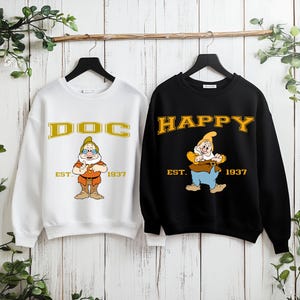 Puede incluir: Dos sudaderas cuelgan de una barra de madera. Una es blanca con "DOC" en letras doradas y un personaje de dibujos animados. La otra es negra con "HAPPY" en letras doradas y un personaje de dibujos animados. Ambas tienen "EST. 1937".