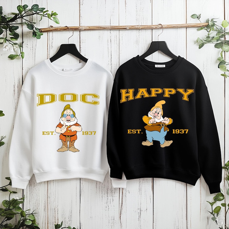 Puede incluir: Dos sudaderas cuelgan de una barra de madera. Una es blanca con "DOC" en letras doradas y un personaje de dibujos animados. La otra es negra con "HAPPY" en letras doradas y un personaje de dibujos animados. Ambas tienen "EST. 1937".