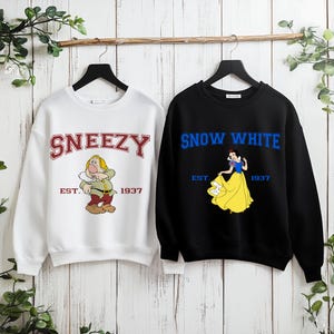 Puede incluir: Dos sudaderas colgadas sobre un fondo de madera. Una es blanca con "Sneezy" en letras rojas y un personaje de dibujos animados. La otra es negra con "Snow White" en letras azules y un gr&aacute;fico de Blancanieves. Ambas tienen "Est. 1937".