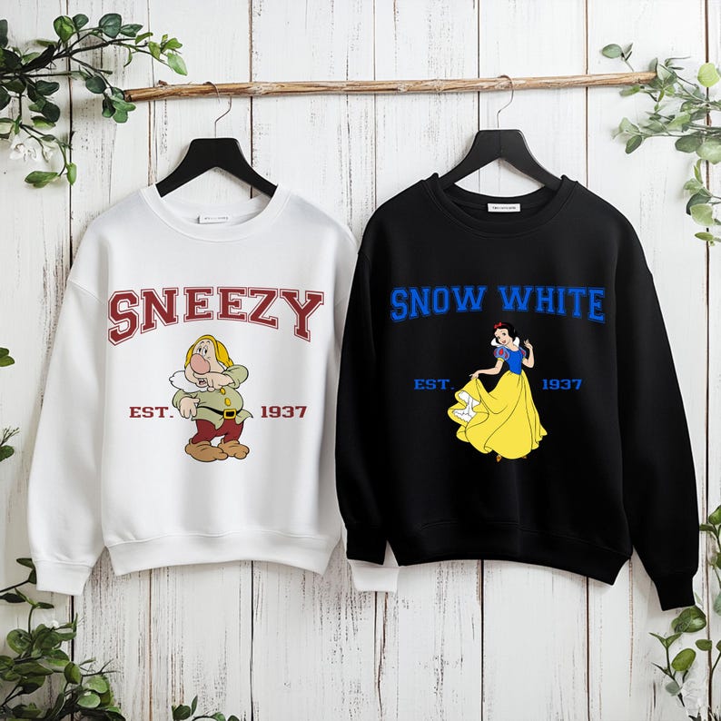 Puede incluir: Dos sudaderas colgadas sobre un fondo de madera. Una es blanca con "Sneezy" en letras rojas y un personaje de dibujos animados. La otra es negra con "Snow White" en letras azules y un gr&aacute;fico de Blancanieves. Ambas tienen "Est. 1937".