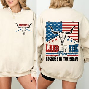 Könnte beinhalten: Cremefarbenes Sweatshirt mit patriotischem Design. Die Vorderseite zeigt das Wort "AMERIKA" über einem Stierschädel und Sternen. Die Rückseite zeigt ein amerikanisches Flaggen-Design mit dem Text "LAND OF THE FREE BECAUSE OF THE BRAVE".