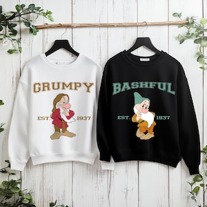 Puede incluir: Dos sudaderas colgando de una barra de madera. Una es blanca con la palabra "GRUMPY" en letras doradas y una imagen de dibujos animados de Gru&ntilde;&oacute;n. La otra es negra con "BASHFUL" en letras verdes y una imagen de dibujos animados de T&iacute;mido. Ambas tienen "EST. 1937".