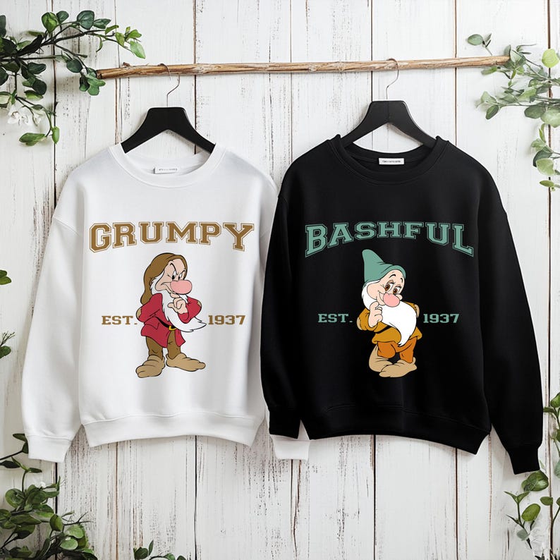 Puede incluir: Dos sudaderas colgando de una barra de madera. Una es blanca con la palabra "GRUMPY" en letras doradas y una imagen de dibujos animados de Gru&ntilde;&oacute;n. La otra es negra con "BASHFUL" en letras verdes y una imagen de dibujos animados de T&iacute;mido. Ambas tienen "EST. 1937".