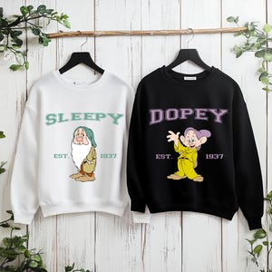 Puede incluir: Dos sudaderas colgadas sobre un fondo de madera. Una es blanca con "SLEEPY" y un gr&aacute;fico de dibujos animados. La otra es negra con "DOPEY" y un gr&aacute;fico de dibujos animados. Ambas tienen "EST. 1937" debajo de los gr&aacute;ficos.