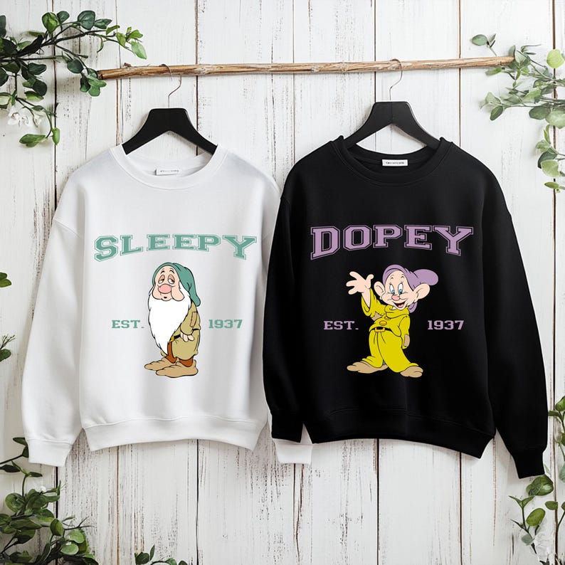 Puede incluir: Dos sudaderas colgadas sobre un fondo de madera. Una es blanca con "SLEEPY" y un gr&aacute;fico de dibujos animados. La otra es negra con "DOPEY" y un gr&aacute;fico de dibujos animados. Ambas tienen "EST. 1937" debajo de los gr&aacute;ficos.