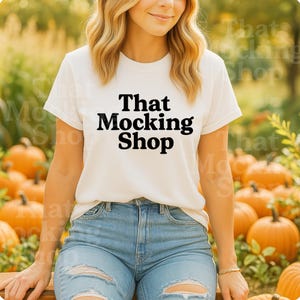 May include: A white t-shirt with the text "That Mocking Shop" in black. The person is wearing light blue ripped jeans. The background features pumpkins and foliage, suggesting an autumn theme.