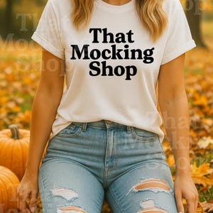 May include: White t-shirt with the text "That Mocking Shop" in black, paired with distressed blue jeans. The scene is set outdoors with pumpkins and autumn leaves.