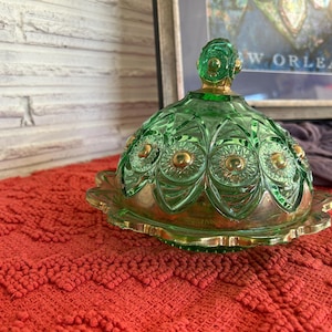 May include: A green glass butter dish with a domed lid and gold-coloured accents. The dish sits on a red textured surface. The lid has a decorative knob on top.