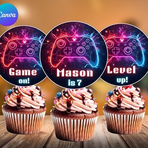May include: Three cupcakes with toppers featuring a neon-style video game controller design. The toppers have text that reads "Game on!", "Mason is 7", and "Level up!". The cupcakes have chocolate frosting and are decorated with sprinkles and chocolate drizzle.