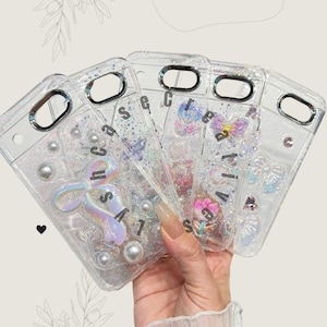 May include: Clear phone cases with glitter, pearls, and decorative elements. One case features a holographic bow and the word "secret". Other cases have butterfly and floral designs. The cases are held in a hand.