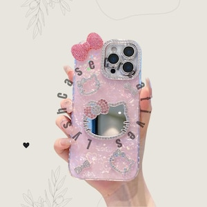 May include: Pink phone case with a Hello Kitty design. The case features a bow, rhinestones, and the text "shacase". The case has a glittery finish and is decorated with Hello Kitty faces.