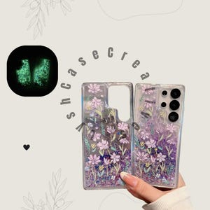 Może przedstawiać: Przezroczyste etui na telefon z kwiatowym wzorem. Etui mają różowe i fioletowe kwiaty z brokatowymi akcentami. Etui są trzymane na jasnym tle. Na tle widnieje napis "shcasecrea".