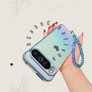May include: Iridescent phone case with the word "creative" and a heart symbol. The case has a blue beaded wrist strap. The phone case is held in a hand.