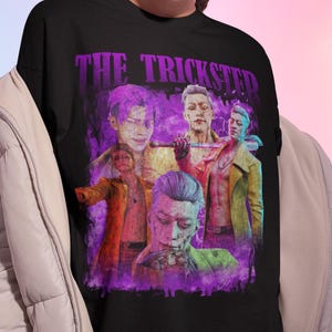 May include: Black t-shirt with a graphic design featuring the text "THE TRICKSTER" in purple. The design includes multiple figures in various poses, with a purple and orange colour scheme. The shirt is worn by a person.
