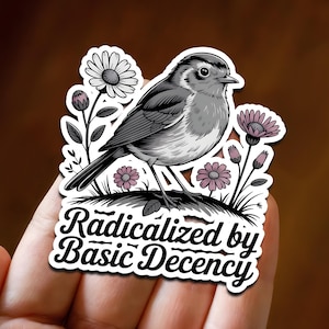 May include: A sticker featuring a detailed illustration of a bird perched among flowers. The bird is in shades of gray and white, with a black outline. The sticker includes the text "Radicalized by Basic Decency" in a stylized font.