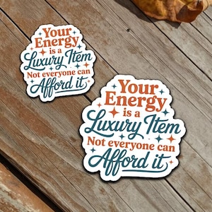 Puede incluir: Dos pegatinas con el texto "Your Energy is a Luxury Item. Not everyone can Afford it." Las pegatinas tienen un fondo blanco con un borde turquesa y texto naranja y turquesa. Las pegatinas están sobre una superficie de madera con hojas secas.