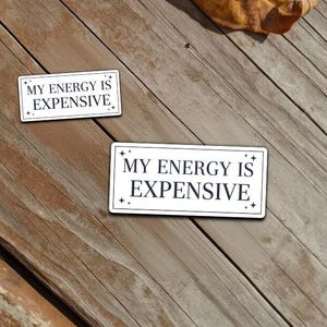 Könnte beinhalten: Zwei rechteckige weiße Emaille-Anstecknadeln mit dem Text "MY ENERGY IS EXPENSIVE" in schwarzer Schrift. Die Anstecknadeln liegen auf einer verwitterten Holzoberfläche mit verstreuten, getrockneten Blättern.