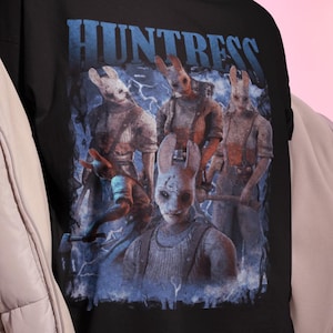 May include: Black t-shirt with a graphic of the Huntress from Dead by Daylight. The design includes the word "HUNTRESS" in blue, and images of the character with a rabbit mask. The shirt is worn with a light beige puffer jacket.