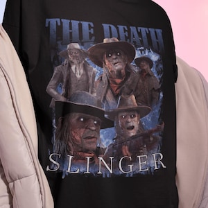 May include: Black t-shirt with the text "THE DEATH SLINGER" in white and blue, featuring a graphic of four figures in hats and period clothing. The shirt is worn by a person.