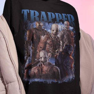May include: Black t-shirt with the word "TRAPPED" in blue block letters. The shirt features a graphic of several figures with masks and menacing expressions. The figures are in shades of brown, grey, and blue.