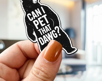 May include: A black and white sticker of a bear with the text "CAN I PET THAT DAWG?". The bear has a white outline and is in a standing pose. The sticker is held by a hand with brown nail polish.