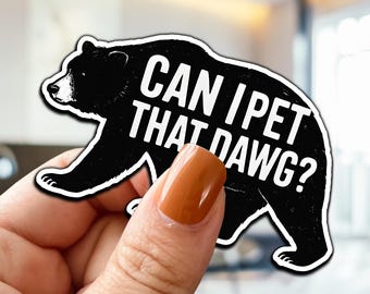 May include: Black bear sticker with the text "CAN I PET THAT DAWG?" in white. The bear is walking to the left with a white outline. The sticker is held by a hand with a brown nail polish.