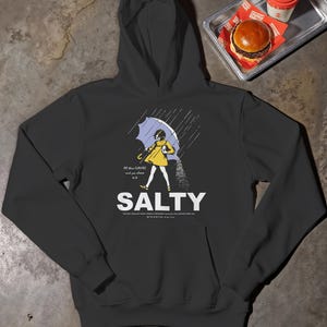 K&ouml;nnte beinhalten: Dunkelgrauer Kapuzenpullover mit einem Front-Grafik einer Person unter einem Regenschirm, die Salz streut. Das Wort "SALTY" steht unter der Grafik, mit zus&auml;tzlichem Text dar&uuml;ber und darunter. Ein Burger und ein Getr&auml;nk sind im Hintergrund zu sehen.