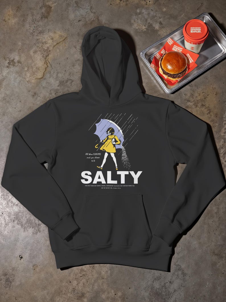 K&ouml;nnte beinhalten: Dunkelgrauer Kapuzenpullover mit einem Front-Grafik einer Person unter einem Regenschirm, die Salz streut. Das Wort "SALTY" steht unter der Grafik, mit zus&auml;tzlichem Text dar&uuml;ber und darunter. Ein Burger und ein Getr&auml;nk sind im Hintergrund zu sehen.