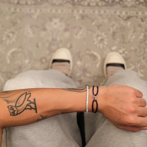Könnte beinhalten: Nahaufnahme von zwei Armbändern an einem Arm. Eines ist ein Tennisarmband mit kleinen, klaren Steinen. Das andere ist ein dunkelblaues Armband mit zwei ovalen Gliedern. Ein Eulen-Tattoo ist auf dem Arm zu sehen.