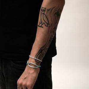 Könnte beinhalten: Arm einer Person mit einem schwarzen Tattoo einer Eule und abstrakten Designs. Die Person trägt zwei Armbänder: eines mit kleinen, klaren Steinen und ein hellblaues Armband.