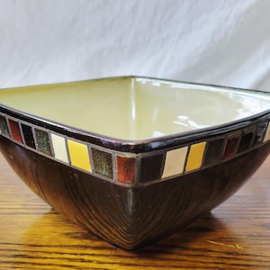 Peut inclure: Bol en céramique carré avec un extérieur marron foncé et un intérieur jaune clair. Le bord est décoré d'un motif en mosaïque de carreaux marron, rouges, jaunes et blancs. Le bol est posé sur une surface en bois.