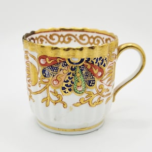 May include: A white teacup with a gold rim and handle, featuring ornate gold, red, blue, and green floral patterns. The cup has a scalloped edge and is likely vintage or antique.