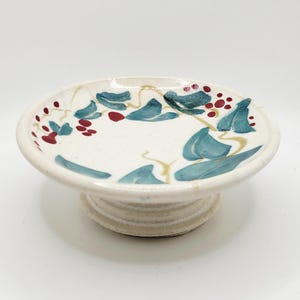 Puede incluir: Un pequeño plato de cerámica blanquecina sobre pedestal con un diseño pintado a mano. El diseño presenta hojas de color verde azulado y bayas rojas, con vides doradas. El plato tiene una parte superior redonda y una base corta y acanalada.