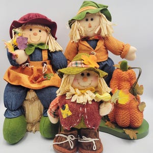 May include: Three plush scarecrow dolls with straw hair, fabric clothing, and hats. One doll wears a maroon hat, one a green hat, and one a smaller green hat. They are accompanied by a textured orange fabric pumpkin with green leaves.