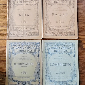 May include: Four vintage opera librettos with text in Italian, English, and German. The covers are beige and blue, featuring titles like "Aida" and "Faust" with the Oliver Ditson Company logo. The books are arranged on a wooden surface.