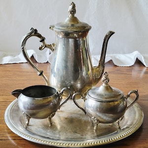 May include: A silver-plated tea service set consisting of a tall teapot with a lid, a creamer, and a lidded sugar bowl, all resting on a round silver-plated serving tray. Each piece features ornate, curved handles and decorative feet.