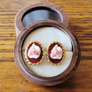 Peut inclure: Boucles d'oreilles à clous en forme de rose, présentées dans un écrin en bois. Les boucles d'oreilles présentent une rose rose sur un fond rouge foncé, bordée d'une bordure dorée en forme de corde. L'écrin est ouvert, révélant un intérieur clair.