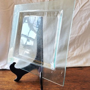 May include: A clear, square glass plate with a textured surface, displayed on a black metal stand. The plate has a slightly raised inner square and a textured border. The background is a light-colored fabric and a wooden surface.