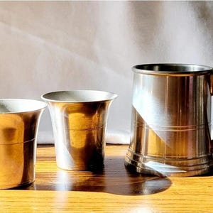 Puede incluir: Tres recipientes metálicos para beber sobre una superficie de madera. Dos son pequeñas copas acampanadas, y el tercero es una jarra grande con asa. Los recipientes tienen un acabado metálico brillante, que refleja la luz.